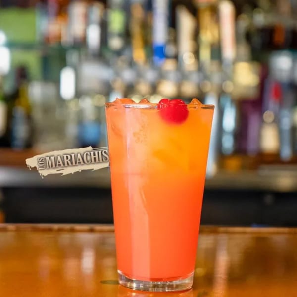 A vibrant orange and red cocktail with a cherry, branded "Los Mariachis", on a bar.