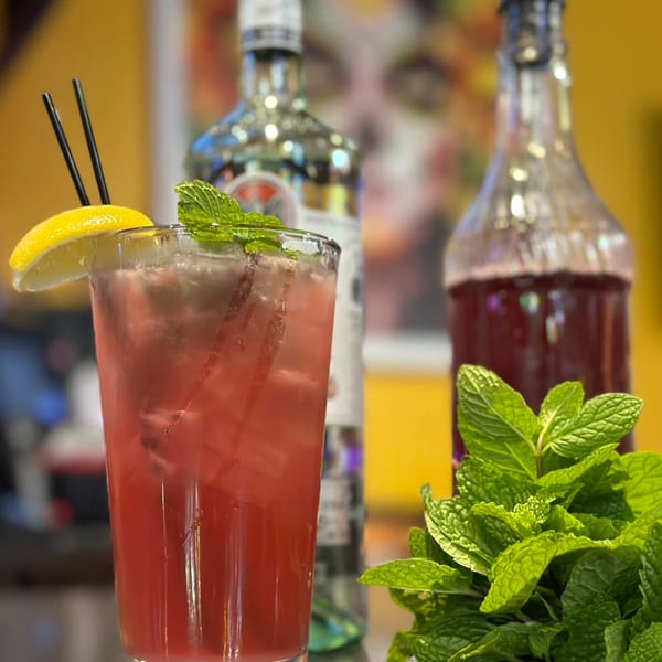 Red cocktail with lemon, mint, and straws, with bottles and mint sprigs in the background.