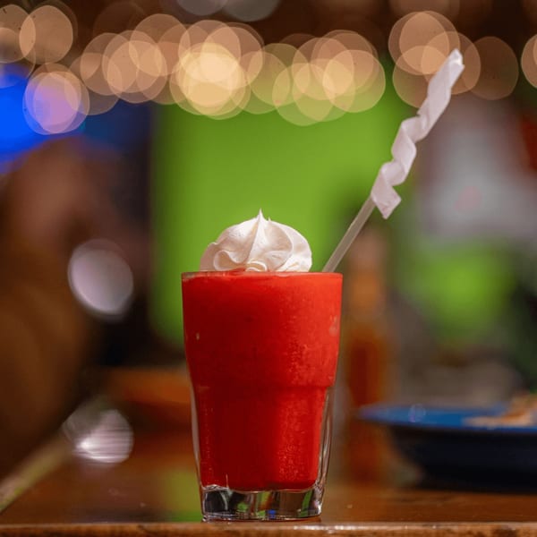 A vibrant red frozen drink topped with whipped cream and a straw, against a blurred background of lights.