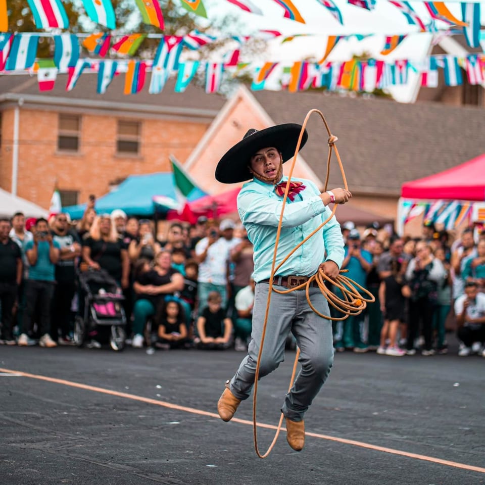 A man in a sombrero and light blue shirt jumps while twirling a rope at a Mexican festival.