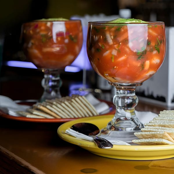 Two large glasses of ceviche with avocado and crackers on yellow and orange plates.