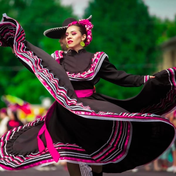 A woman in a black and pink traditional Mexican dress dances, with a man in a sombrero behind her.