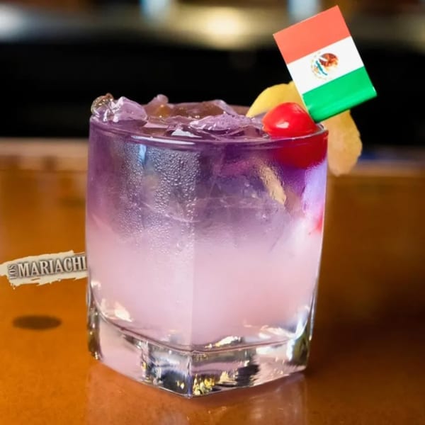 Purple cocktail with ice, a cherry, lemon, and a Mexican flag pick on a bar.