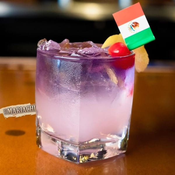 Purple cocktail with ice, a cherry, lemon slice, and a Mexican flag pick on a bar.