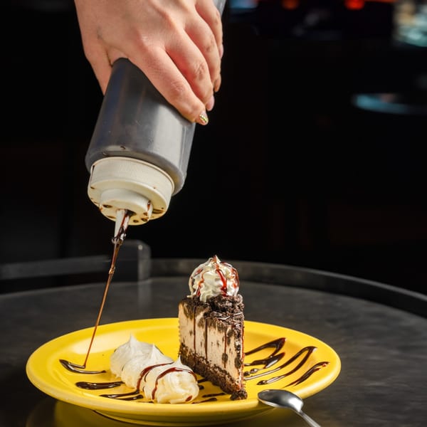 A hand drizzles chocolate syrup onto a slice of cookies and cream cheesecake with whipped cream.